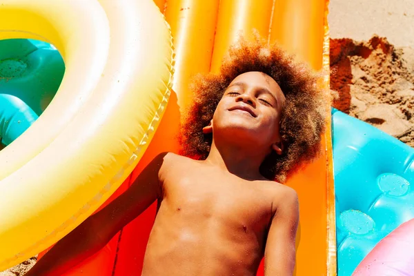 Above view portrait of African boy enjoying summertime, laying on big heap of swimming mattresses with his eyes closed — Stock Image View Portrait African Boy Enjoying Summertime Laying Big Heap Swimming Royalty Free Stock Photos