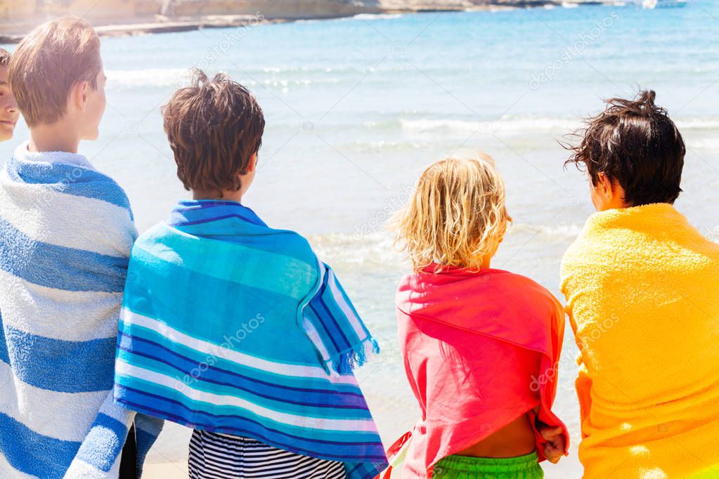 Back view portrait of four age-diverse boys wrapped in beach towels looking at summer sea