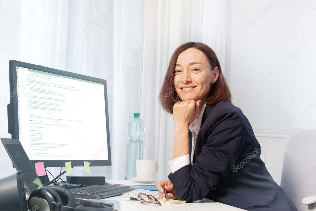 Beautiful middle aged woman in business suit working with computer, sitting at the desk in the office