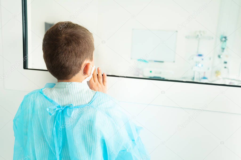 Little boy in hospital robe wearing medical mask worried pointing finger near glass of operating room