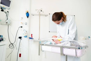Hastanede maske ve bornoz giyen hemşire yoğun bakımünitesinde prematüre bebek bebeğini elinde tutuyor