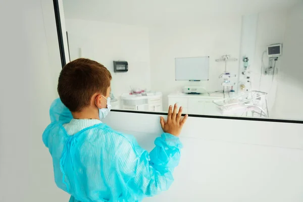 医療マスクとローブを着て病院のIcu部屋の窓の近くに小さな男の子 — ストック写真