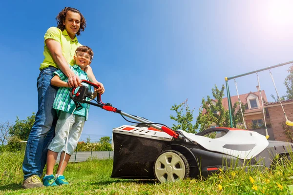 裏庭で電動芝刈り機を使って芝生を掘る息子を教える若い父親の低角の肖像画 — ストック写真
