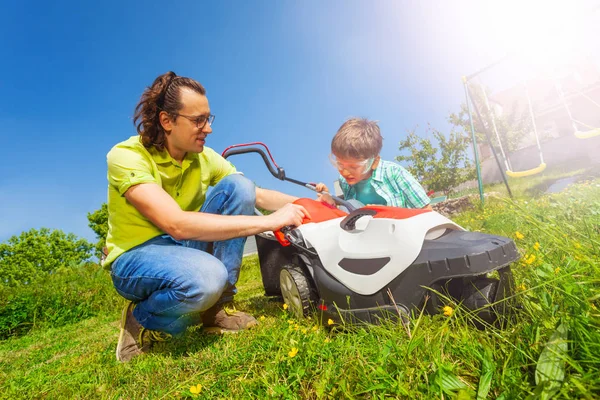 若い父親は 裏庭で芝生を切った後 芝刈り機の草箱をきれいにするために彼の息子を教える — ストック写真
