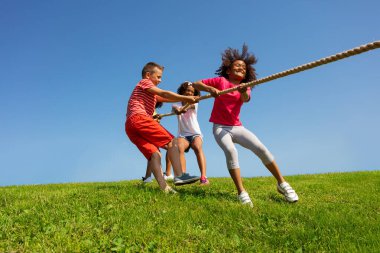 Bir grup çocuk, parktaki mavi gökyüzünde tutkulu ve duygusal bir şekilde halat çekiyorlar.