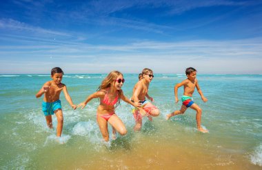 Bir grup mutlu kız ve erkek çocuk denizden el ele tutuşup su sıçratarak, gülerek, eğlenerek koşuyor.