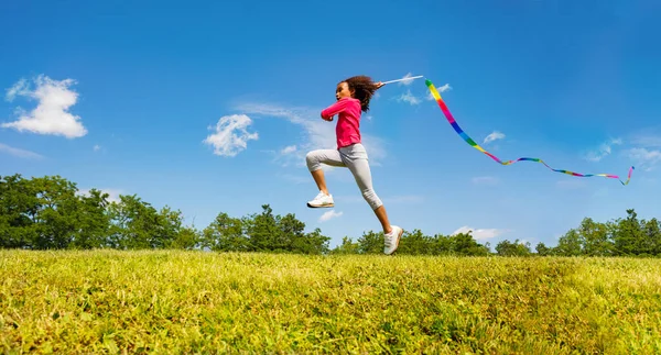 可愛いです女の子長いですジャンプ振っていますカラフルなリボン以上フィールドと空の背景 — ストック写真