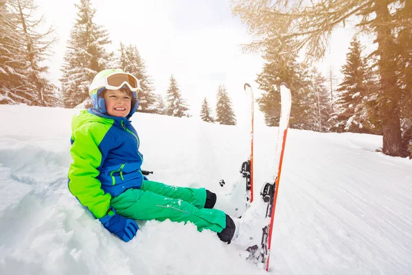 小さな男の子は マスク ヘルメットとスキーの笑顔を着て森の中のパスの近くの雪の中に座っている — ストック写真