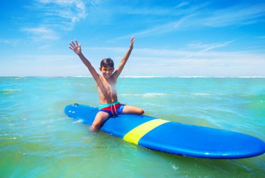 Mutlu küçük çocuk denizde ellerini kaldırmış sörf tahtasına oturuyor.