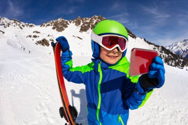 Küçük çocuk ayakta durup alp kayağı kaskını takıyor ve dağdan maske takıyor cep telefonu kamerasıyla selfie çekiyor.