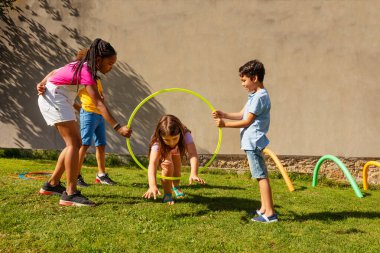 Bir grup arkadaş renkli spor çemberleriyle oynuyorlar, küçük uzun saçlı kız içeride iki çocuğun ambarına atlıyor.