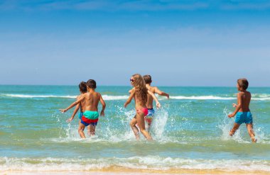 Bir grup çocuk sahile birlikte su sıçratarak dalgalara doğru koşuyor.