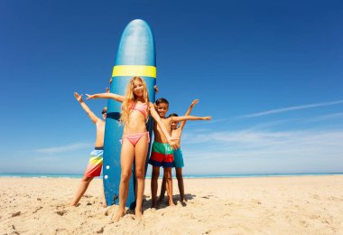 Küçük güzel kız sörf tahtasında durup bir grup arkadaşla el sallıyor.