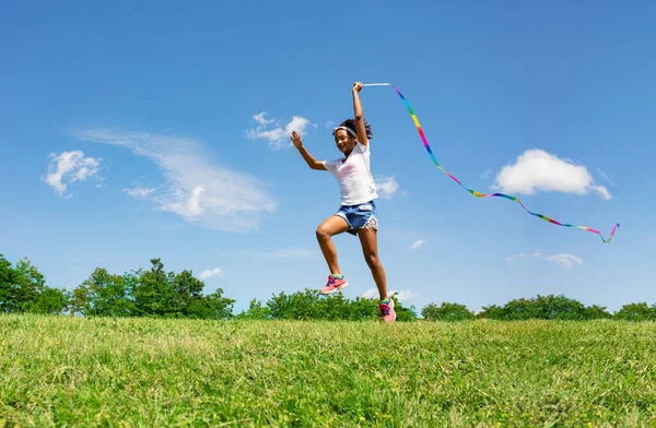可爱的小女孩在公园里挥动着彩带在田野和天空的背景上奔跑 — 图库照片