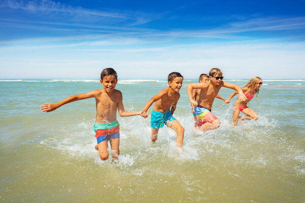 Group of cute boys and girls friends run together holding hands from sea making splashes, laughing, having fun