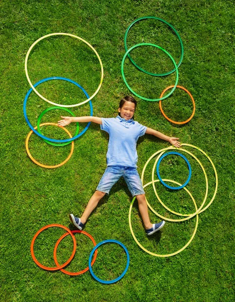 小白种人快乐的小男孩躺在草地上 地上挂着许多五彩斑斓的呼啦圈 从上面看他的画像 — 图库照片