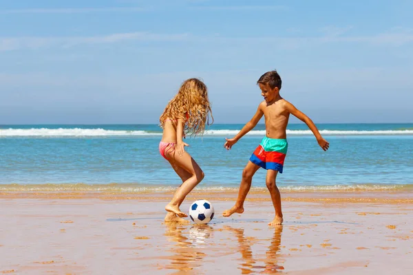 Two kids boy and girl play soccer football ball on sand beach near the wate...