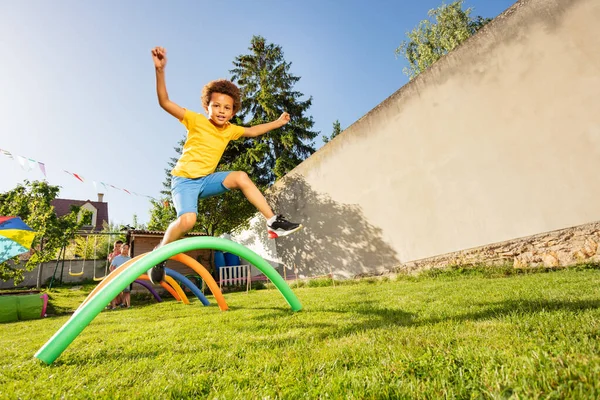Happy black boy jump over sequence of obstacles on the backyard playground with his hands up smiling