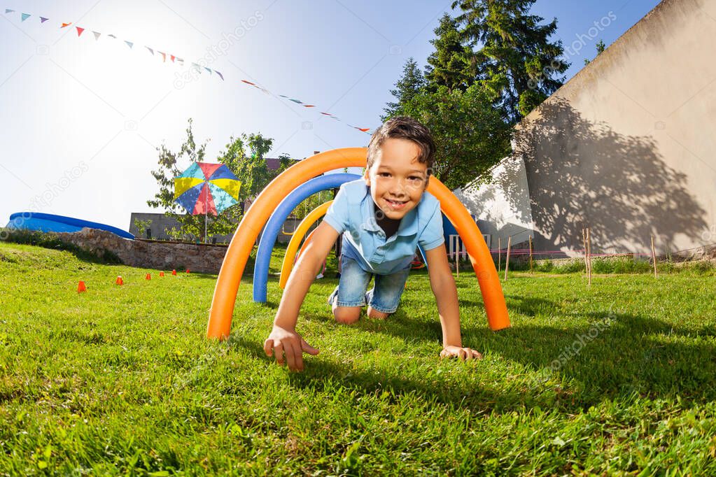 Handsome Caucasian boy pass obstacle course tunnel on his fours on playground lawn