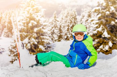 Kar maskesi ve miğfer takmış çocuk portresi beyaz köknar ağaçlarının üzerinde kışın karda oturur.