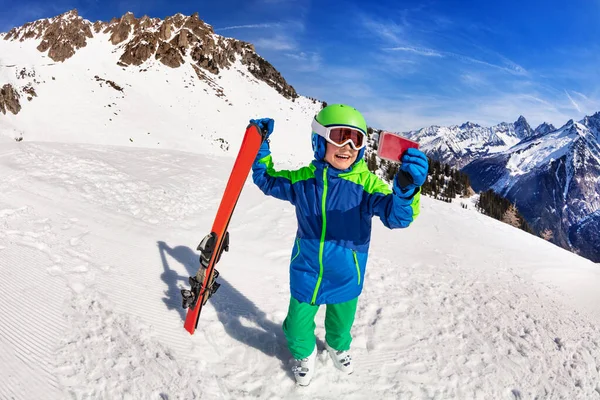 携帯電話のカメラで写真を撮る小さな男の子は山の上に巨大な高山スキーウェアヘルメットとマスクスタンド — ストック写真