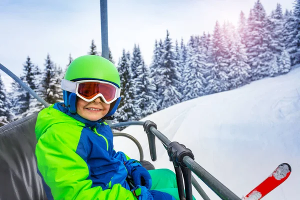 スキーウェアのヘルメットとマスクを持つ少年の近くの肖像画は 山の斜面の上に笑みを浮かべてケーブルスキルの椅子に座っている — ストック写真