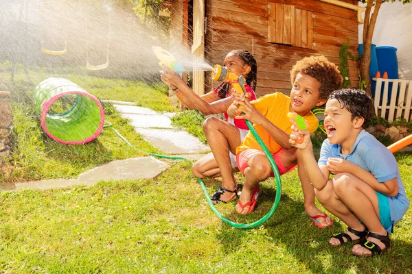 Group of kids squat and shoot with water guns, garden hose playing game on backyard