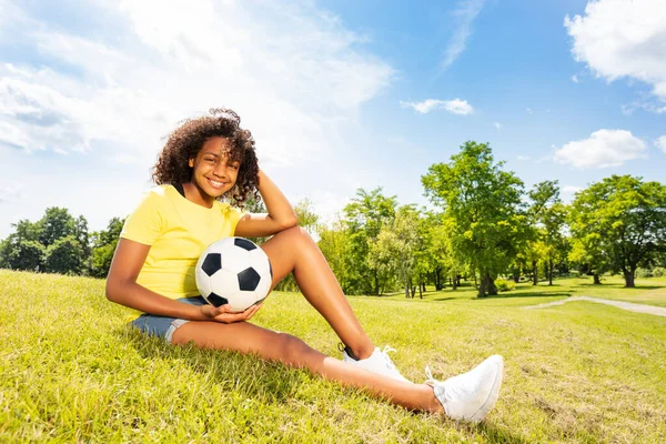 若いです幸せなかわいいです巻き黒美しいです女の子とともにサッカーボール座ってザ草 — ストック写真