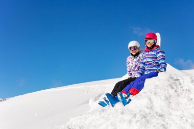 İki tatlı kız mavi arka planda kar yığınının üzerinde oturuyor. Gökyüzü maskesiyle ve kamerada gülümseyen gözlüklerle.