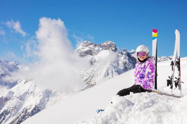 Kayak kıyafetli, kasklı ve maskeli mutlu bir kız dağın tepesinde kar üzerinde oturur kameraya bakar.