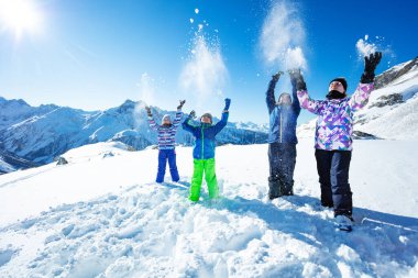 Dört mutlu kayakçı çocuk dağın tepesinde dikilirken havaya kar fırlatıyor.