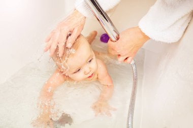 Anne banyosunda küçük bir çocuk banyo küvetinde duş başlığını sıcak suyla yıkıyor.