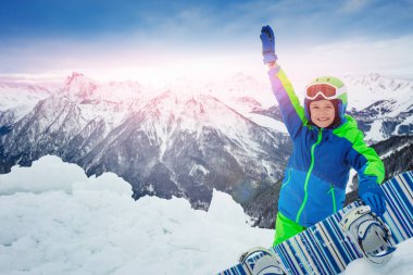 Snowboard 'la kar kaldıran bir çocuğun yakın portresi arka planda dağ zirvesinde büyük bir gülümseme sergiliyor