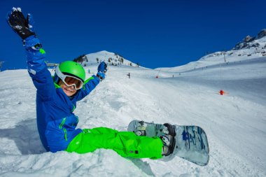 Snowboard yapan küçük bir çocuğun yakın görüntüsü kar manzarasında maske takmış, kayak kaskı kaldırıyor.