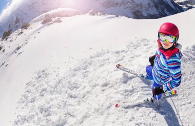 Bir dağın tepesinde kar üstünde kayak yapan bir kızın portresi. Pembe kaskla parlak bir ceket giyiyor.