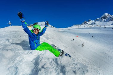Snowboard 'lu küçük çocuk kar manzarasında maske takmış, kayak kaskını kaldırıyor.