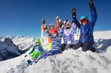 Büyük bir grup kayakçı çocuk renkli spor kıyafetleriyle dağların tepelerinde kar yığınının üzerinde oturur ve ellerini kaldırır.