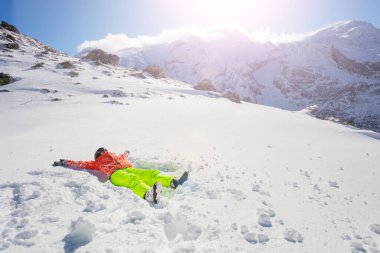 Mutlu genç kız karda uzanırken arka planda dağ olan turuncu sarı kayak kıyafeti giyerken iyi eğlenceler.