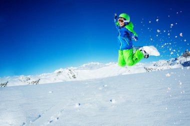 Mutlu çocuk mavi gökyüzü ve dağın tepesinde kar tozu ile elleri kaldırarak snowboard 'a atlıyor.