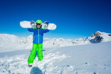 Omuzlarında snowboard tutan bir çocuğun mutlu tam boy portresi. Arkaplanda dağın tepesindeki manzaranın üzerinde duruyor.
