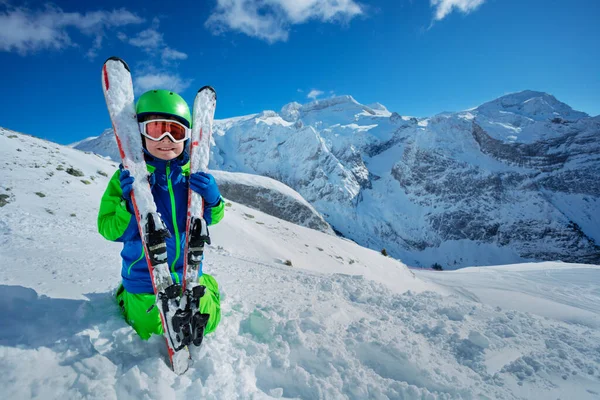 Happy smiling boy in mask and green helmet hugging skis sitting in snow over mountain ridge