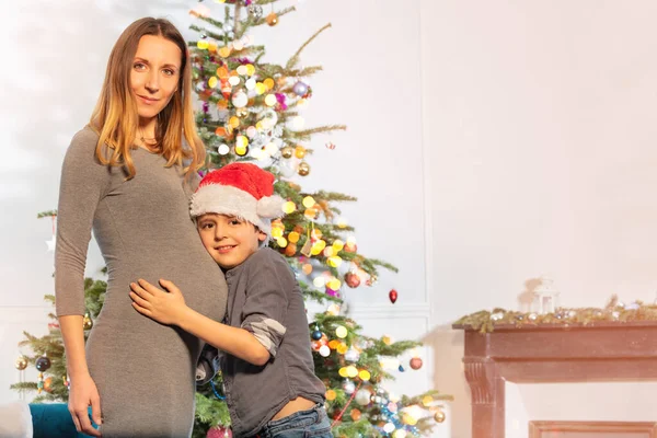 靠近圣诞树的妈妈和小男孩头戴圣诞帽抱着妈妈的肚子的画像 — 图库照片