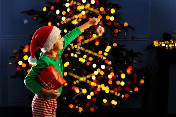 小男孩用手指指了指 站在那里 把圣诞礼物放在点着灯的圣诞树旁 — 图库照片