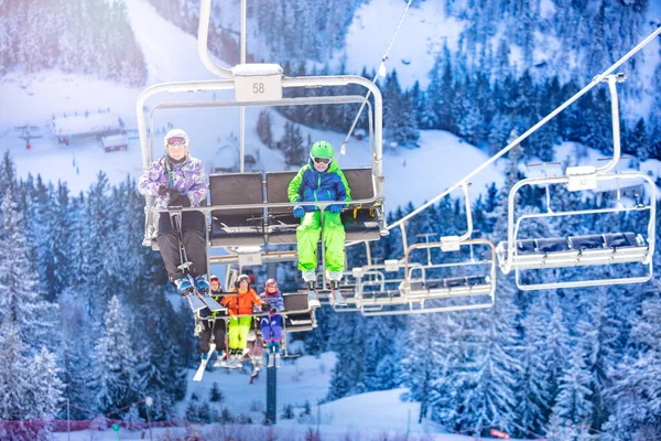 Boy and girl sit on chairlift with friends going on second chair all kids in colorful outfit over snow forest on background