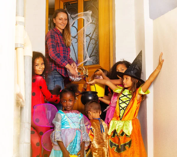 一緒に家のドアの近くに立っている女性からお菓子を受け取る衣装で子供たちのグループとハロウィンライフスタイルシーン — ストック写真