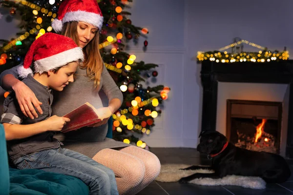 怀孕的母亲和她的儿子坐在圣诞树旁和壁炉边看圣诞童话故事 — 图库照片