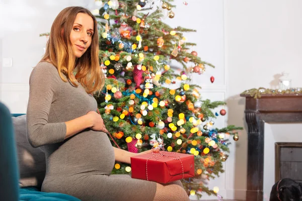 孕妇摸着肚子 坐在靠近圣诞树的沙发上 拿着礼物 — 图库照片