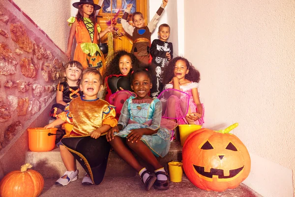 衣装を着たハロウィーンの子供たちの大グループは オレンジのカボチャとバケツの笑顔で階段に座ってカメラを見ています — ストック写真