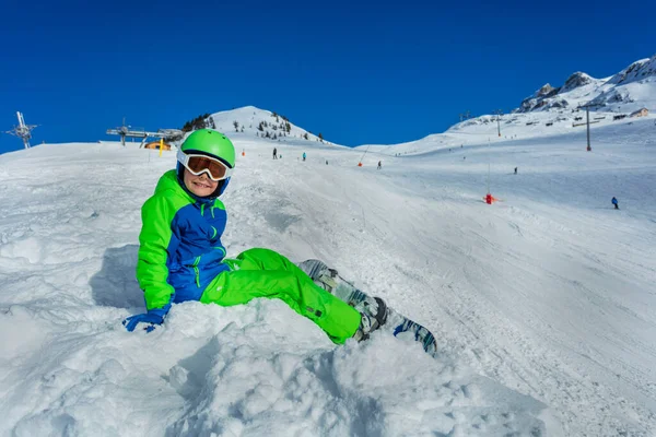 スノーボードを持つ小さな男の子がマスクの側から雪景色に座って スキーヘルメット笑顔 — ストック写真