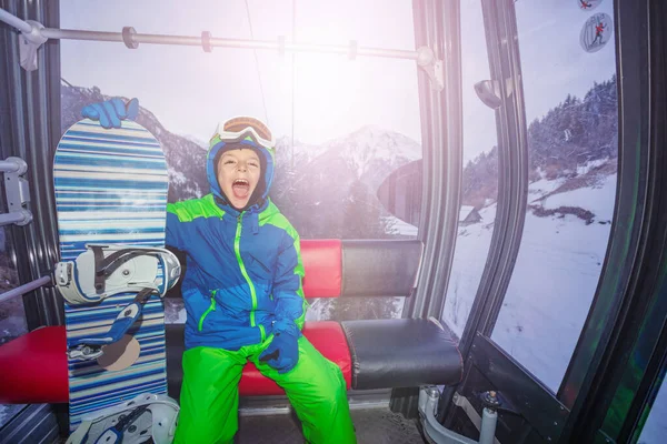 冬のリゾートのスキーリフトキャビンに座っている幸せから叫んでスノーボードを持つ幸せな少年 — ストック写真